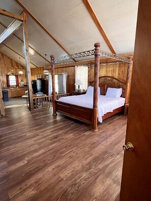Una cama con dosel de madera en una habitación con suelo de madera y una cocina al fondo.