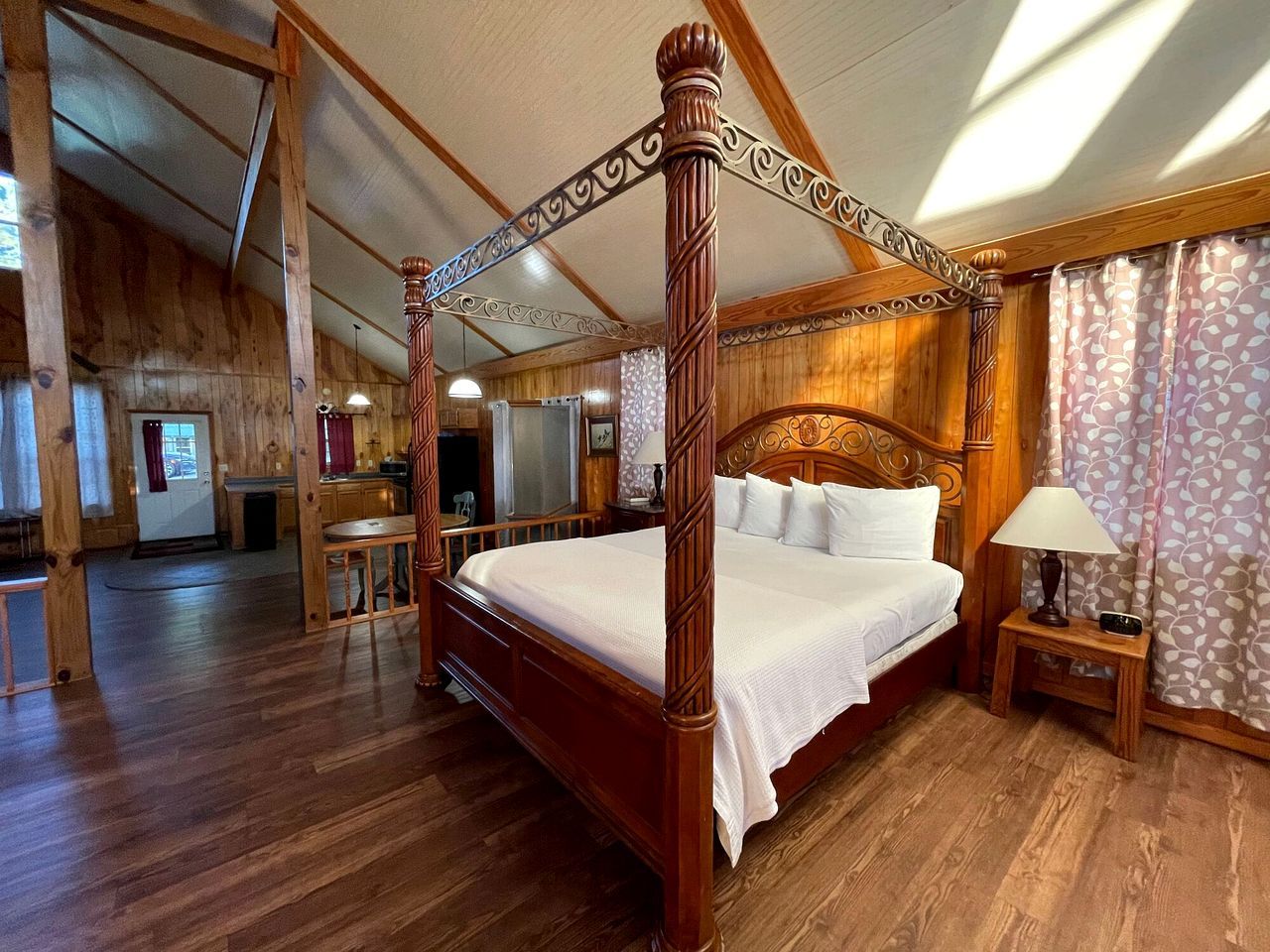 A four-poster bed with a wooden headboard, a bedside table with a lamp, and a view of a kitchen area.
