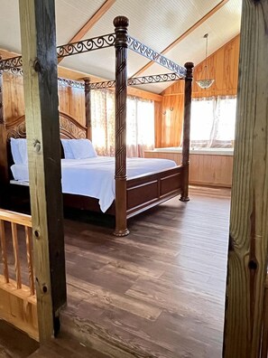 A four-poster bed with a wooden headboard and footboard, a wooden floor, and a window with curtains.