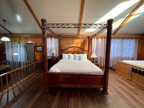 A four-poster bed with a wooden frame and white bedding, a wooden floor, a wooden ceiling with beams, a small bedside table with a lamp, and a bathroom with a bathtub and a mirror.