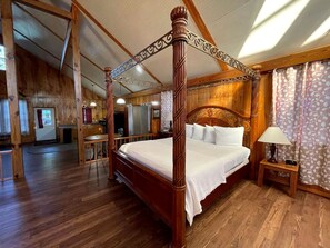 A four-poster bed with a wooden headboard, a bedside table with a lamp, and a view of a kitchen area.