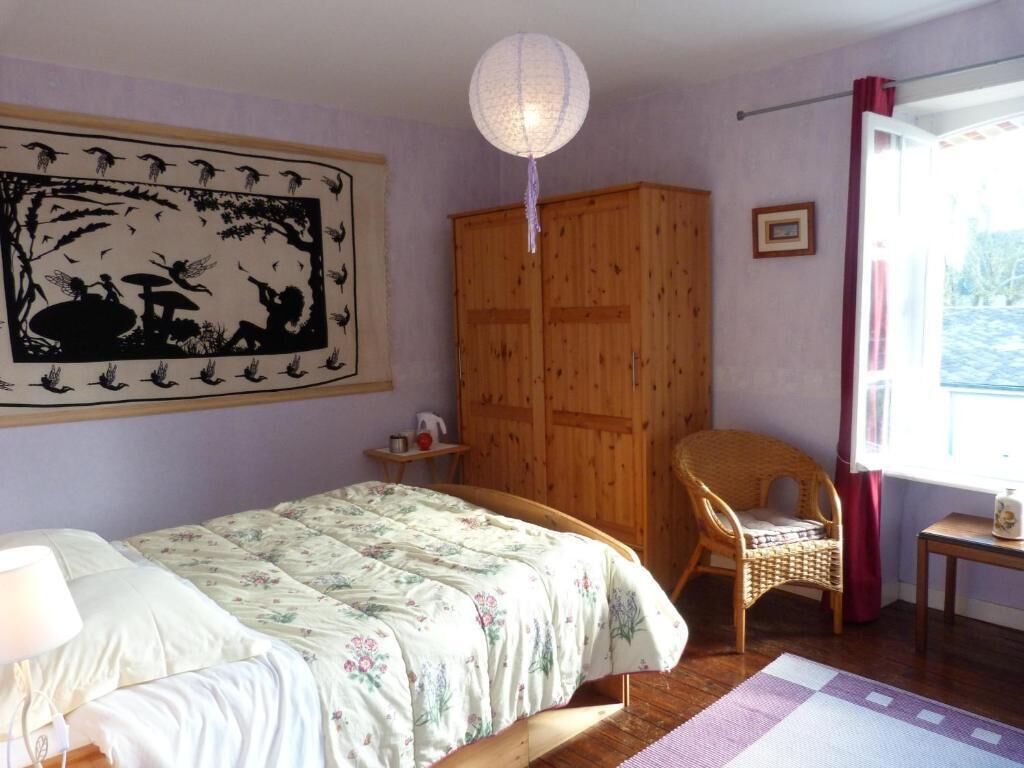 A bedroom with a bed, a wooden wardrobe, a wicker chair, a table, and a hanging light fixture.