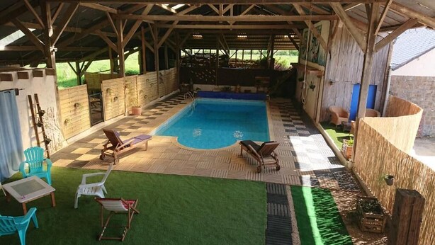 Una piscina exterior con suelo a cuadros, rodeada por una terraza de madera y sillas, bajo una zona techada con vistas al exterior.