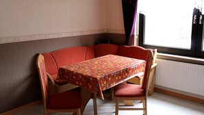 Zona de menjador a la cantonada, amb una taula, caires, un radiador i una finestra amb cortines.