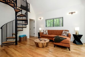 A modern living room with a wooden staircase, a sofa, a coffee table, and a window with blinds. - Cozy and Secluded Cottage in the Woods of Western North Carolina, near Pisgah National Forest (Balsam Grove)