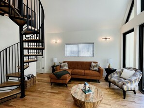 Un salon moderne avec un escalier en colimaçon, un canapé d’angle, un fauteuil à motifs, une table basse et une fenêtre avec des stores.