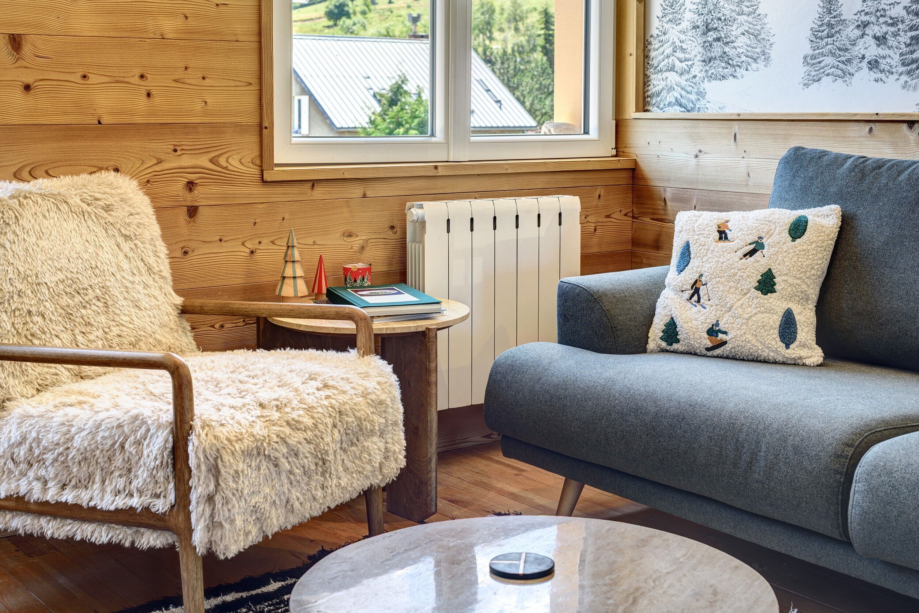 A cozy room with a wooden paneled wall, a fluffy chair, a grey sofa, a small round table, and a radiator.