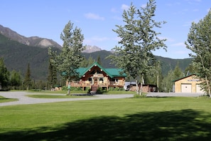 A log cabin with a green roof, surrounded by a well-maintained lawn and trees, with mountains in the background.