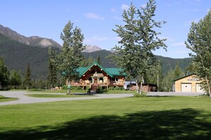 A log cabin with a green roof, surrounded by a well-maintained lawn and trees, with mountains in the background.