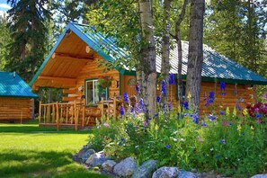 A log cabin with a porch, surrounded by a garden with various flowers and trees.