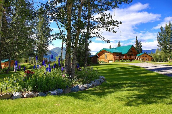 A log cabin with a green roof, surrounded by a well-maintained lawn and flowering plants.