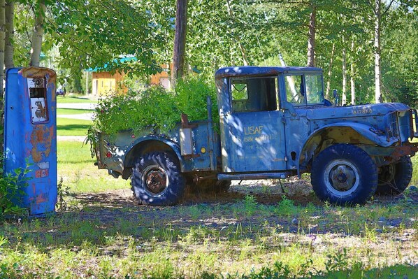 Un veicolo USAF d'epoca con cabina blu parcheggiato accanto a una cassetta utenze blu arrugginita, in un'area erbosa con alberi sullo sfondo.