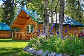 A log cabin with a porch, surrounded by a garden with various flowers and trees.