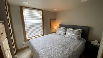 A bedroom with a bed, a nightstand, a lamp, a window with blinds, and a door.