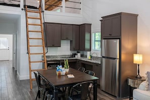 A kitchen with dark cabinets, a stainless steel refrigerator, a wooden ladder, a dining table with chairs, and a small side table with a lamp. - Creek Cabin ADA accessible in Livingston, Texas (Livingston)