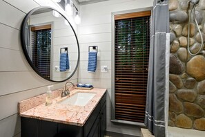 A bathroom with a marble sink, a mirror, a shower with a stone wall, and wooden blinds. - Romantic 1-Bedroom Cabin with Fireplace, Hot Tub & Nature Views Near Broken Bow (Broken Bow)
