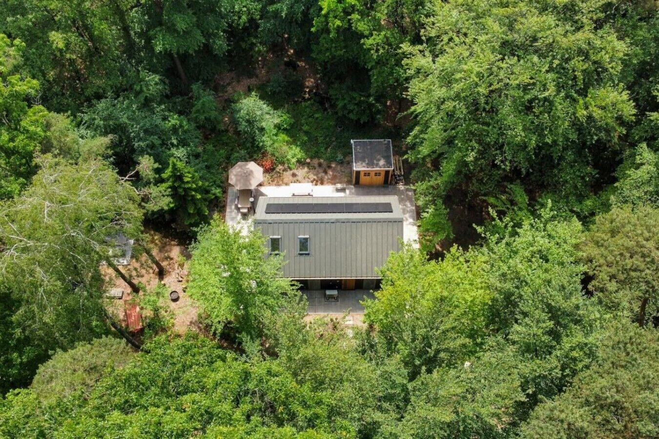 Una casa moderna immersa in una fitta foresta, con un'area patio visibile e un pannello solare sul tetto.