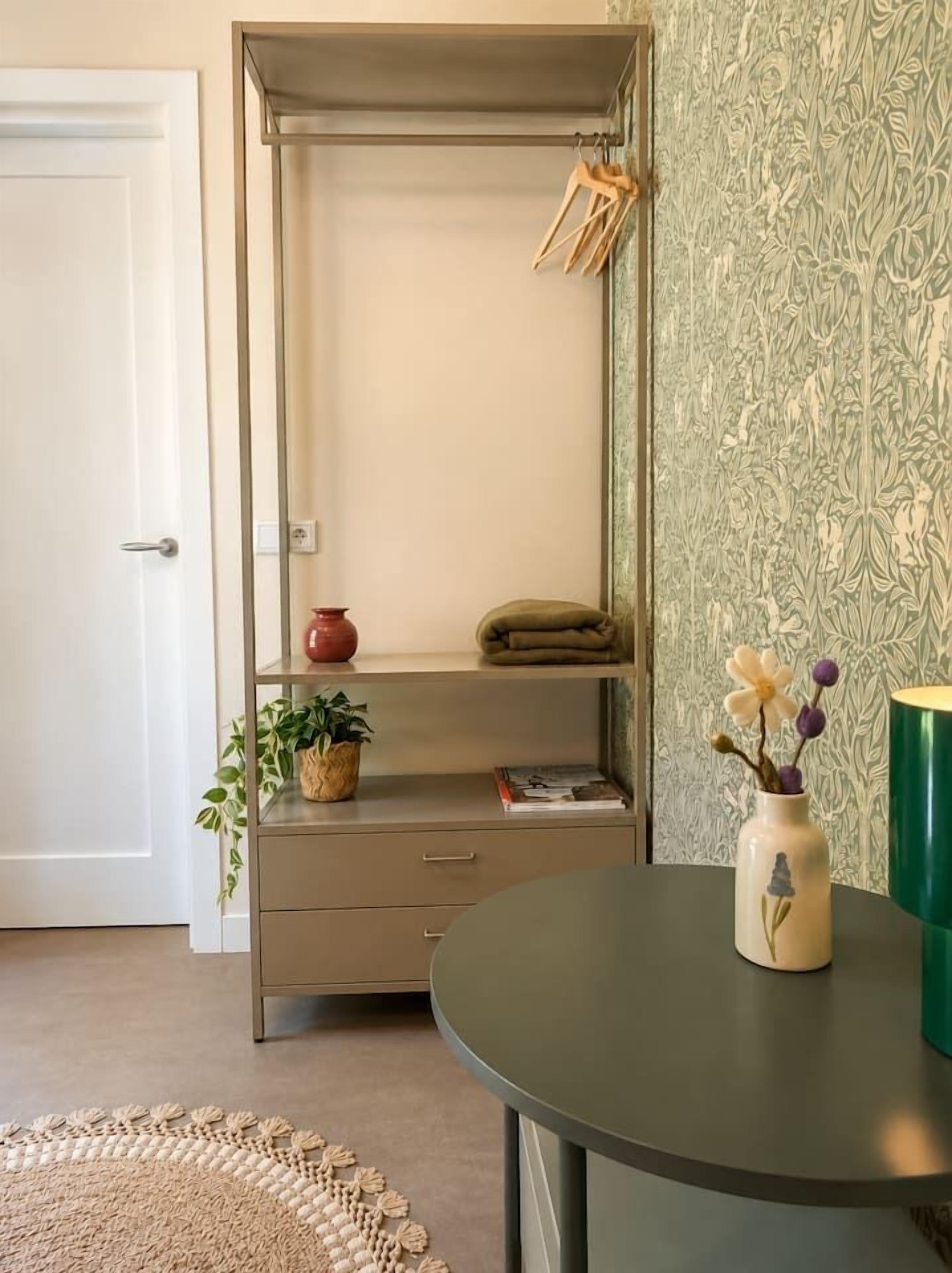 A room with a tall, narrow cabinet with shelves, a round table with a vase, and a patterned wallpaper.