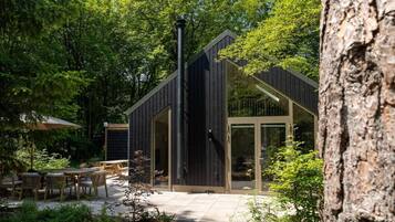 En moderne træhytte med en glasdør og en terrasse med udendørs møbler, omgivet af frodig vegetation.
