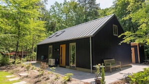 A small, dark-colored building with a sloped roof, a single door, and a small porch area with a bench. - Holiday home Koningsbos – At home in the forest (Emst)