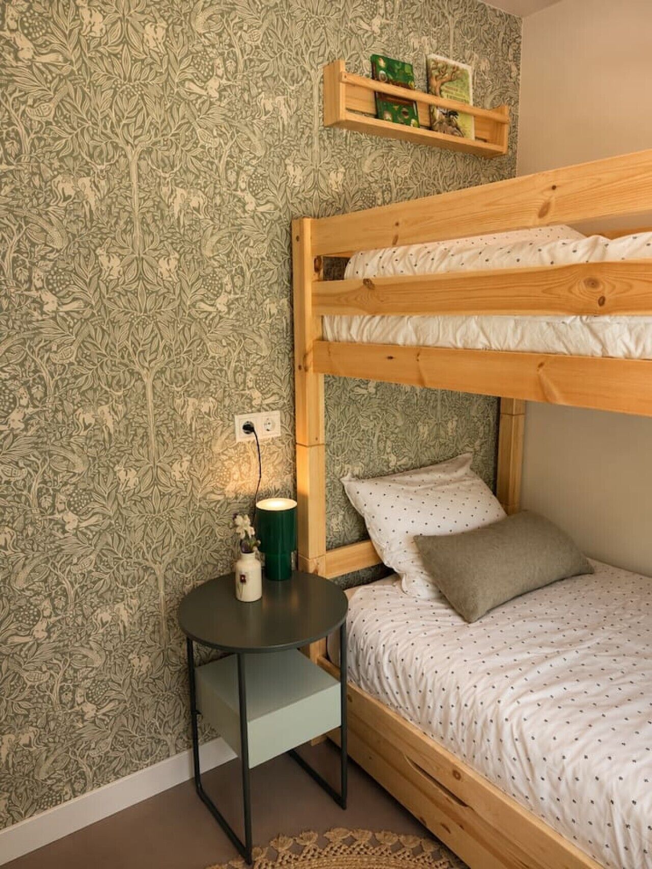 A bunk bed with a side table, a wall-mounted shelf with books, and a patterned wallpaper.