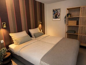 Een keurig opgemaakt bed met hoofdbord, een nachtkastje met een plant en een houten boekenkast met manden.