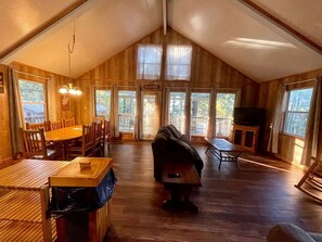 A spacious loft-style room with a high ceiling, wooden floors, a dining area with a table and chairs, a sofa, a fireplace, and large windows. - Inviting Lakefront Cabin with Boat Launch near Zwolle, Louisiana (Zwolle)