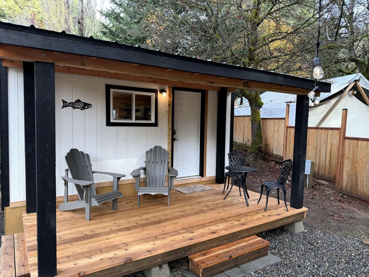 A wooden deck with Adirondack chairs, a table, and a tent in the background.