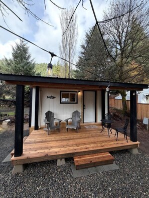 A wooden deck with chairs and a table, a door with a fish sign, and a pergola with a light fixture. - Cute Cozy Cabin for Two for a Romantic Escape in the Umpqua National Forest in Oregon (Idleyld Park)