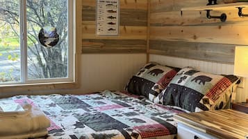 Ein blockhaushaltiger Raum mit einem Bett, einer Steppdecke, einem Fenster, einem an der Wand befestigten Schild und einem Regal mit Gläsern.