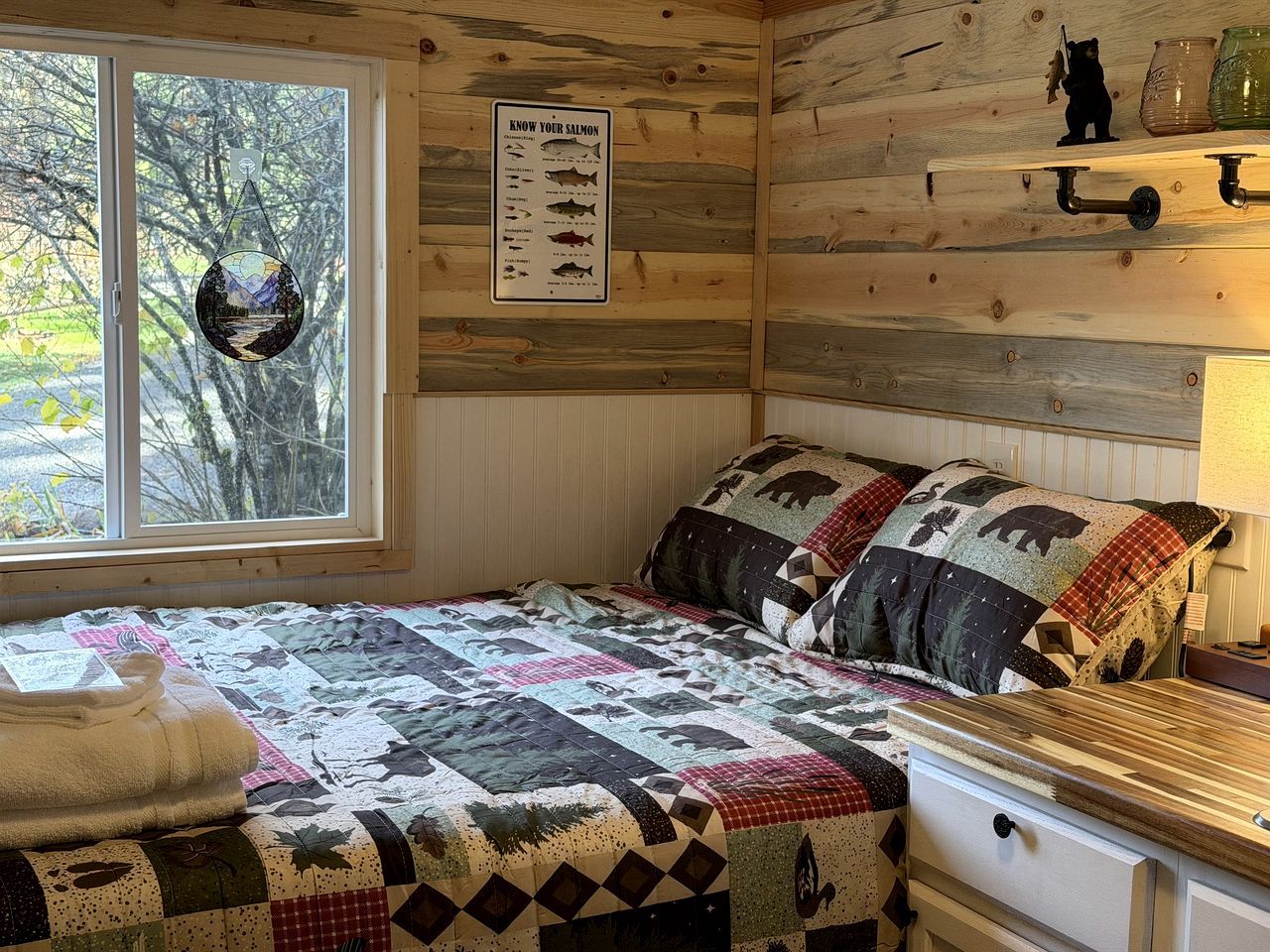 A cabin-style room with a bed, a quilt, a window, a wall-mounted sign, and a shelf with jars.