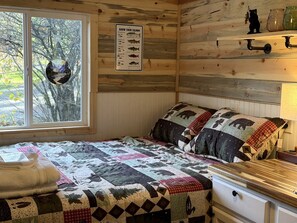 Ein blockhaushaltiger Raum mit einem Bett, einer Steppdecke, einem Fenster, einem an der Wand befestigten Schild und einem Regal mit Gläsern.