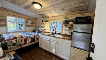 A compact cabin with a kitchenette, a bed with a quilt, and a window with a view of trees.