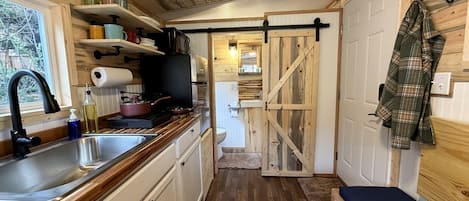 A compact kitchen with a sink, stove, and storage shelves. A sliding door leads to a bathroom with a toilet and sink.