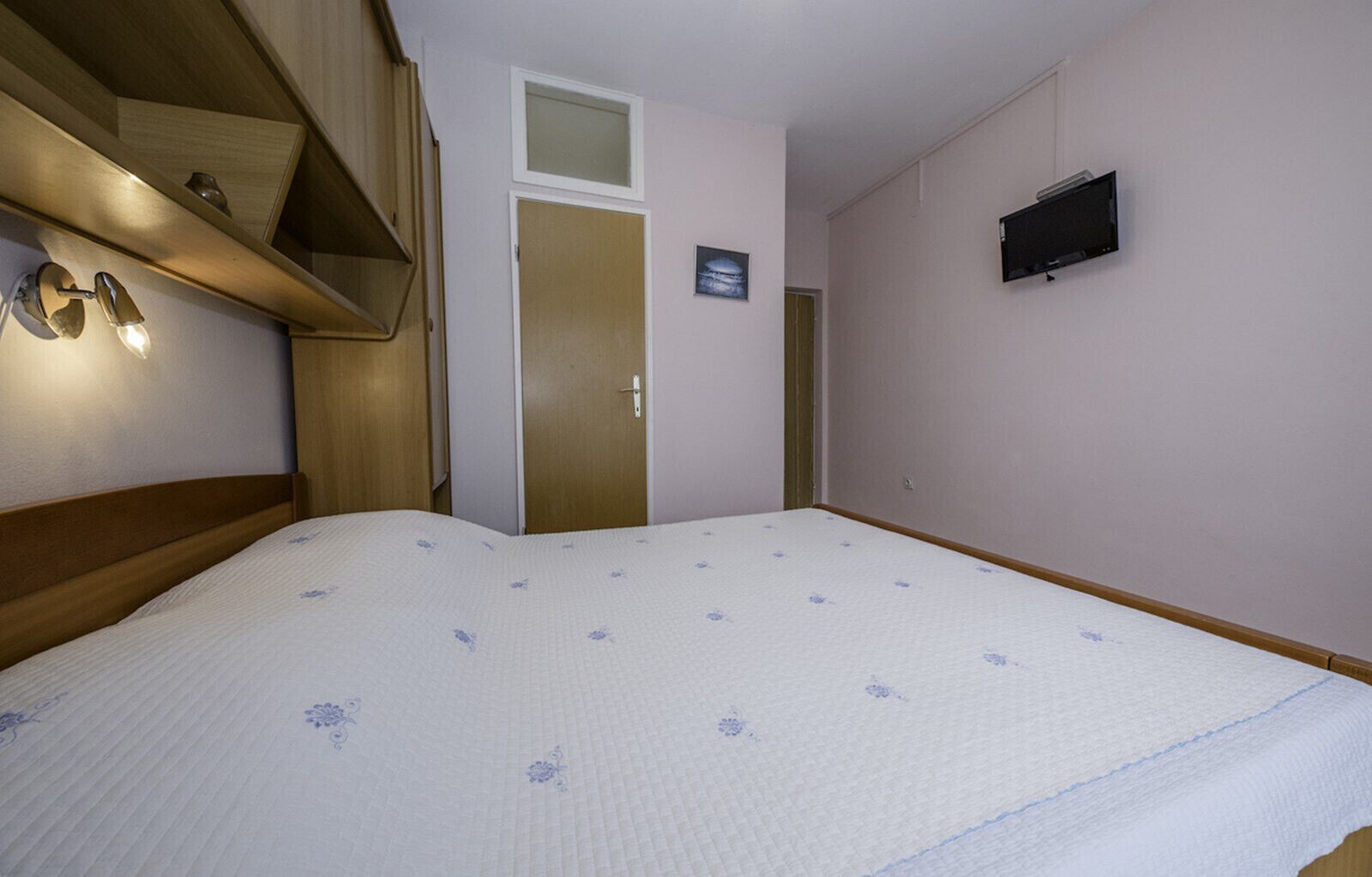 Ein Schlafzimmer mit einem Bett, einem hölzernen Kopfteil, einem an der Wand befestigten Fernseher und einer Tür mit kleinem Fenster.