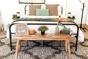 A neatly arranged bedroom with a bed, bedside tables, a bedside lamp, a small table with a sign, a plant, a candle, and books.