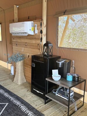 A compact room with a wood-paneled interior, a black coffee machine, a small table with a bowl and mugs, and a window with a view of trees.