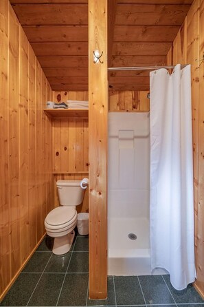 Un baño con paneles de madera, un inodoro, una ducha y un estante sobre el inodoro.