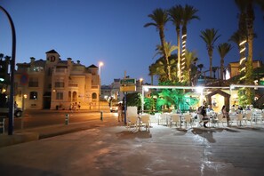 A street scene at dusk with a restaurant patio, palm trees, and a building with a sign reading "Alfred Nobel". - Playa de los Locos Beach Apartment, Torrevieja (Torrevieja)