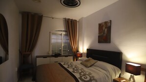 Une chambre d’hôtel avec un lit, des lampes de chevet, une fenêtre avec des rideaux, un tableau au mur et un ventilateur de plafond.