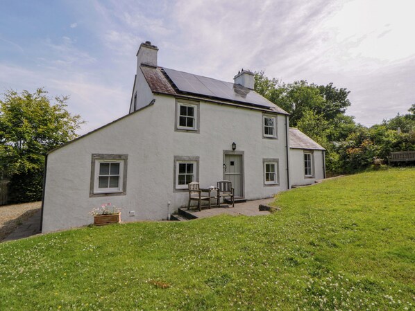 A two-story white house with a sloped roof, a front porch with two chairs, and a small garden area. - Cynefin (Newport)