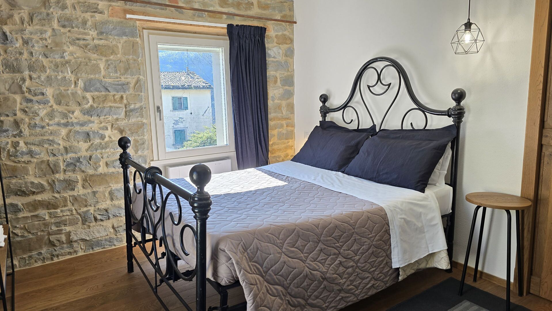 A bed with a quilted cover, a stone wall, a window with curtains, and a hanging light fixture.