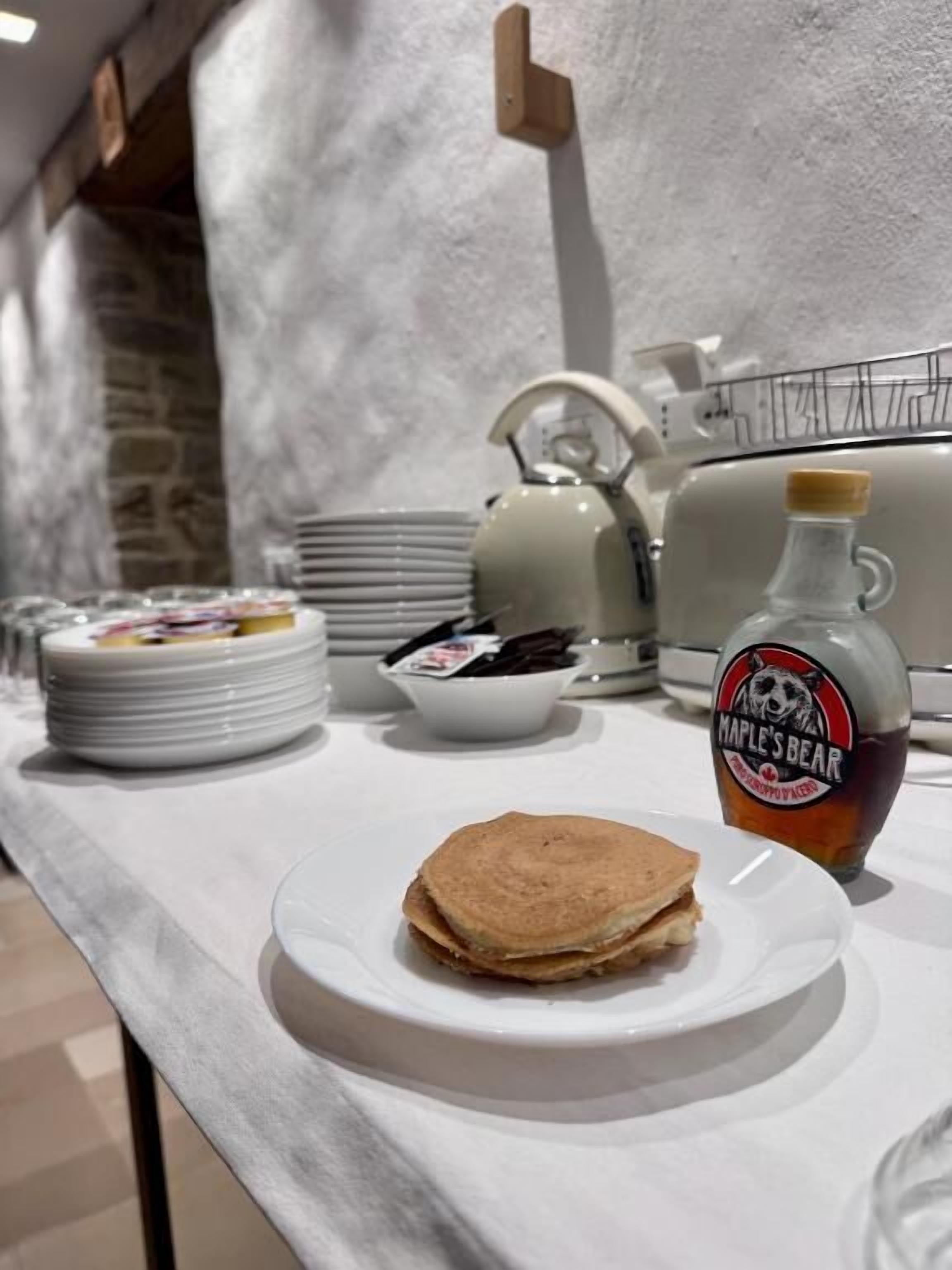 A breakfast setup with pancakes, a bottle of maple syrup, and a kettle on a white table.