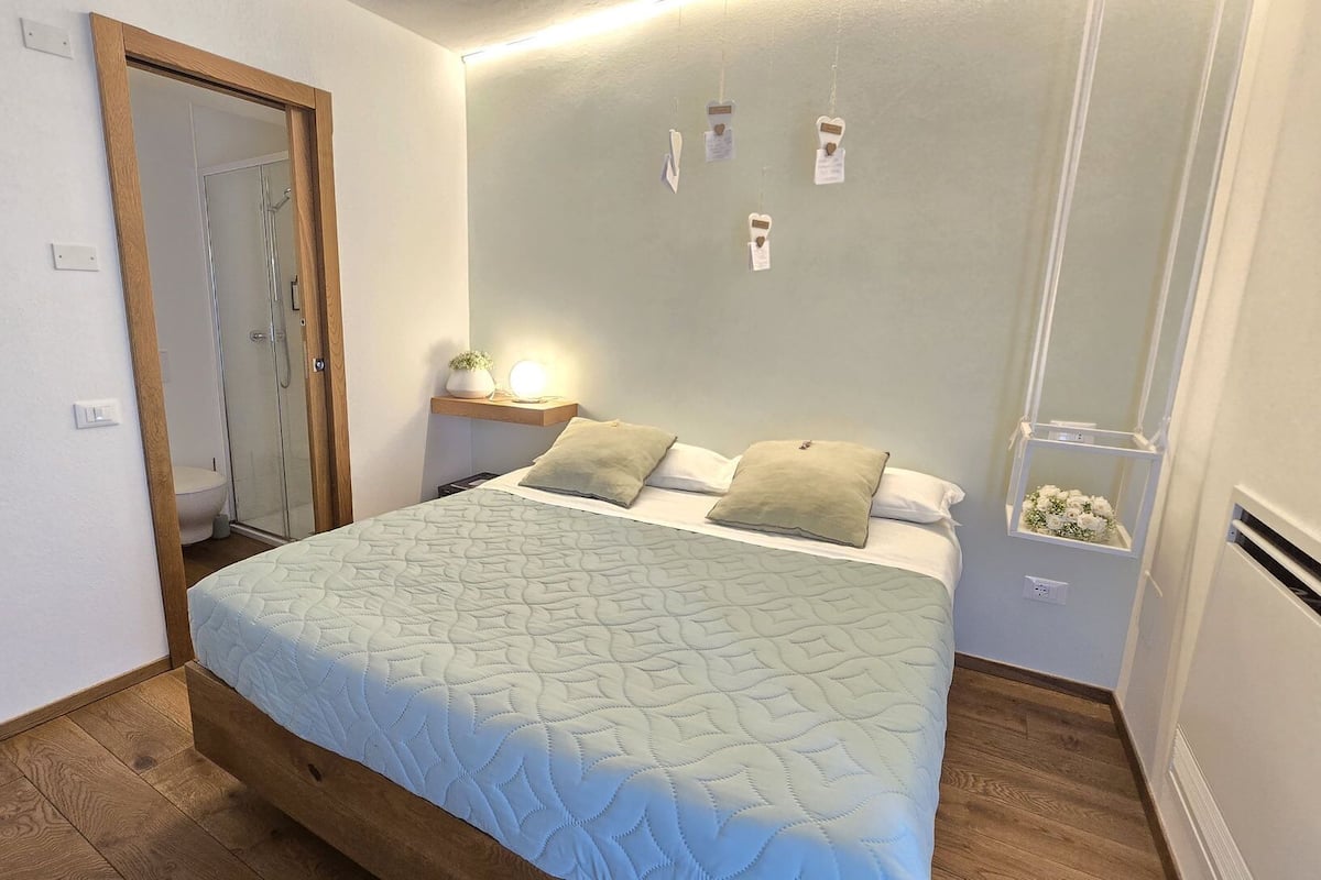 Una camera da letto con un letto, un comodino, uno specchio, una mensola con fiori e una porta che conduce al bagno.