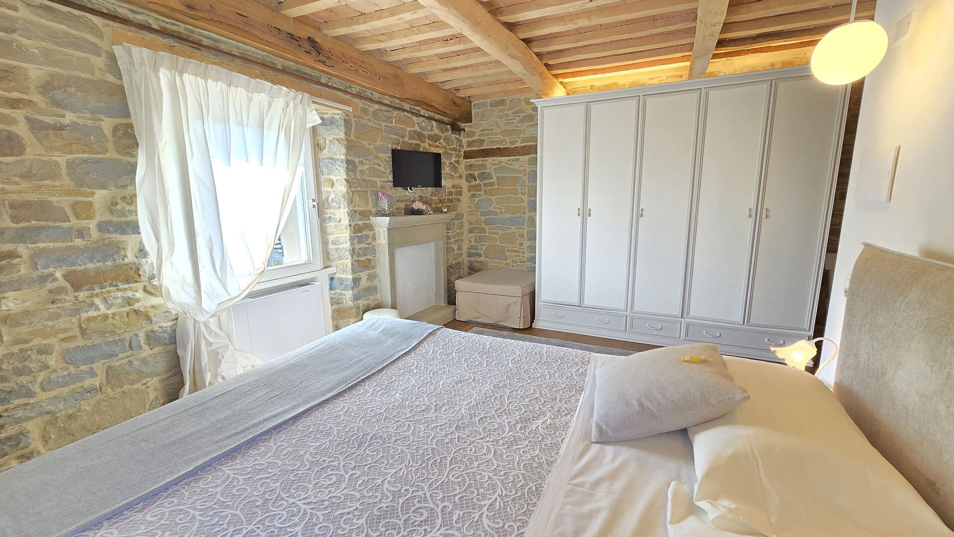 A bedroom with a stone wall, a bed with patterned bedding, a wardrobe, and a television on the wall.