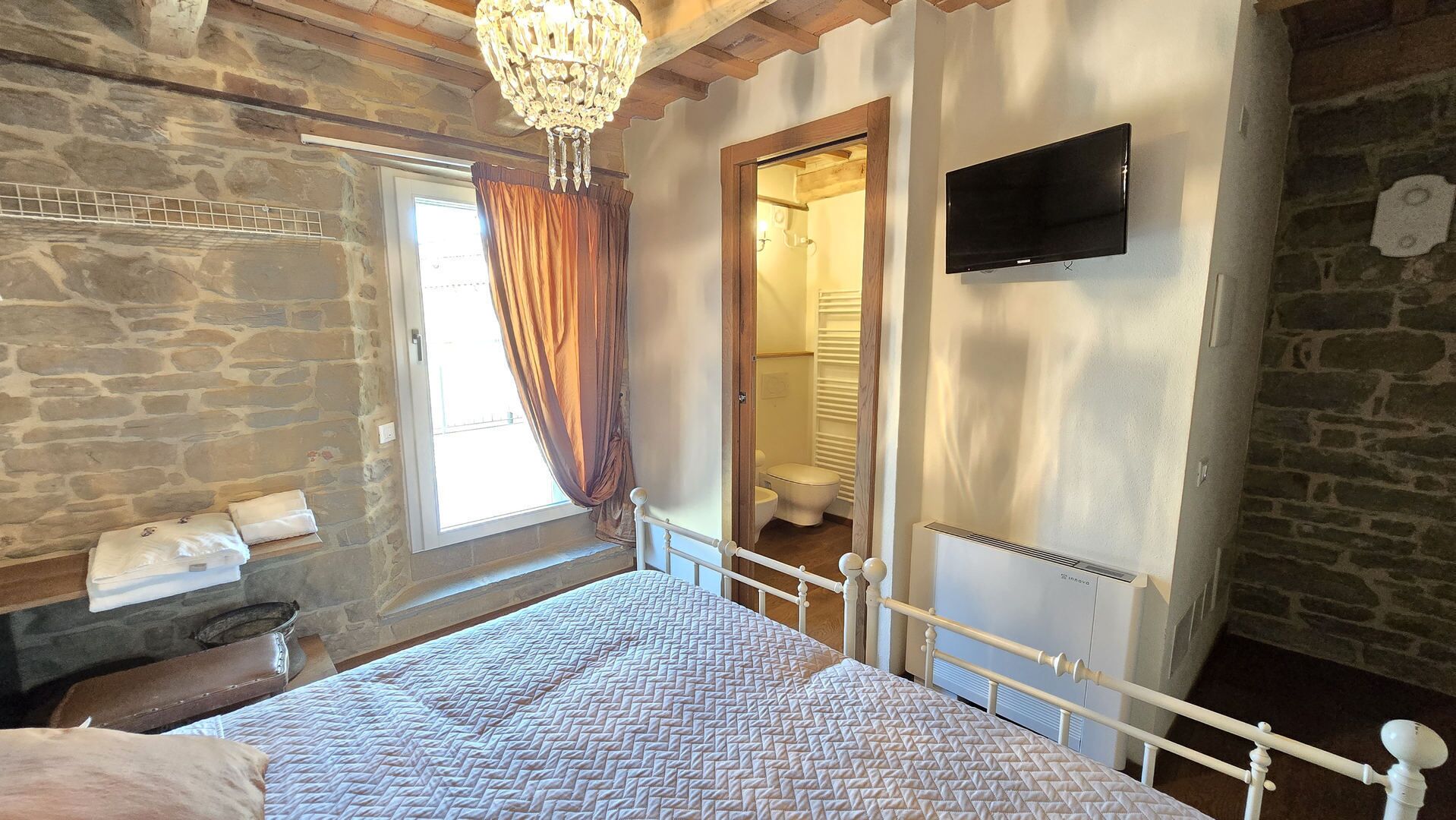 Ein Schlafzimmer mit einem Bett, einem Fernseher, einem Fenster mit Vorhängen, einer Steinwand und einer Holzbalkendecke.