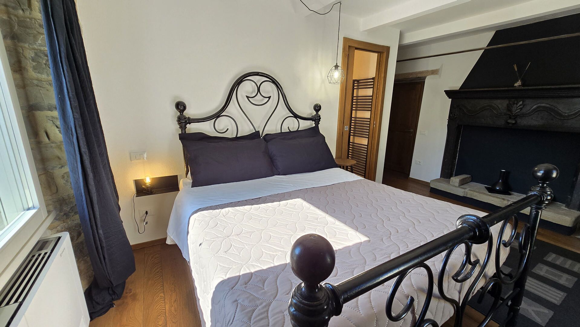 Ein Bett mit Kopfteil, ein Nachttisch mit Lampe, ein Kamin und ein Fenster mit Vorhängen.