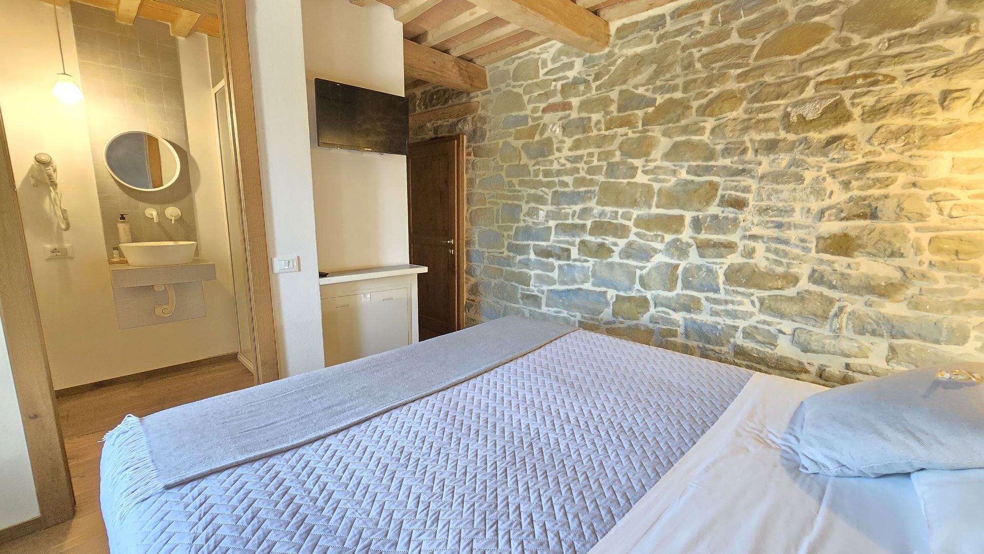Ein Schlafzimmer mit Steinwand, einem Bett mit Quiltdecke, Holzboden und einem Badezimmer mit Waschbecken und Spiegel.