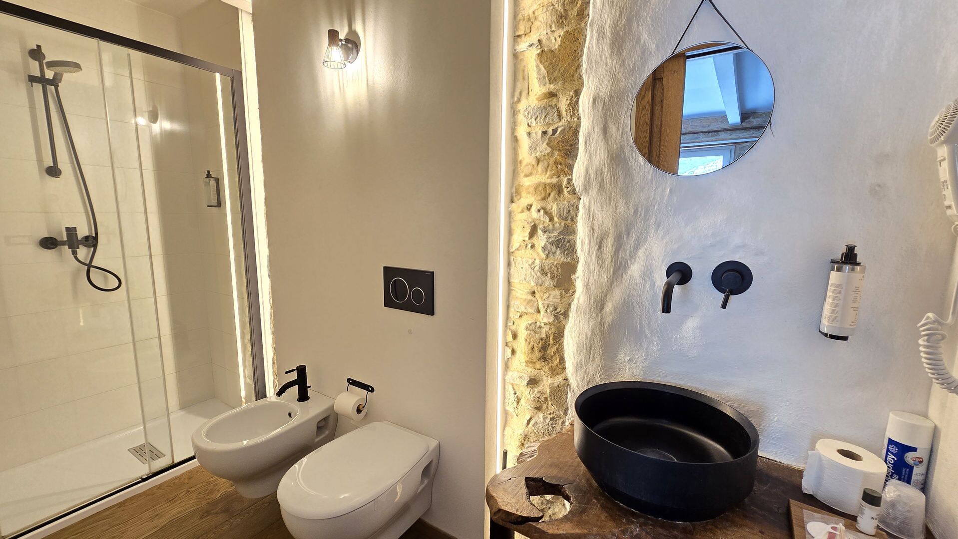 Un baño con ducha, bidé, inodoro y pared de piedra con espejo.