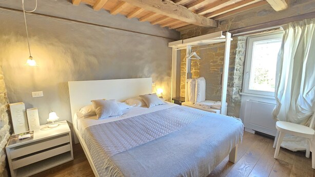 A bedroom with a large bed, bedside tables, a window with curtains, a stone wall, and a wooden ceiling. - Cà Maina (Santa Sofia)